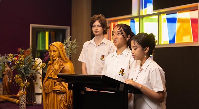 Three St Clare students presenting in the chapel. the background has a Religious statue, and stained glass windows.