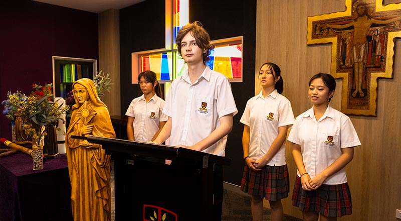Students presenting in Church. There is a cross and stained glass window in the background, with students and a statue of Mary in foreground.