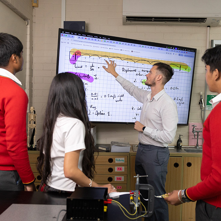 St Clare's Teacher pointing to TV, the TV has maths equations on it.